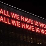 Foto mit großem Neon-Schriftzug und den Sätzen "ALL WE HAVE IS WORKDS" und "ALL WE HABE IS WORLDS"