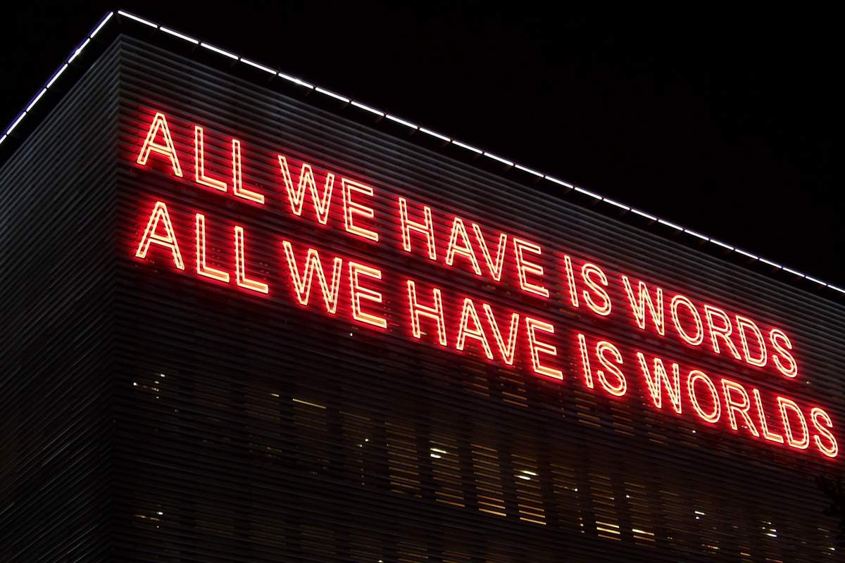 Foto mit großem Neon-Schriftzug und den Sätzen "ALL WE HAVE IS WORKDS" und "ALL WE HABE IS WORLDS"